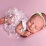 Hallie is registered to the contest to win money with this photo: baby, newborn, sleeping, pink_background, white_dress, floral_dress, headband, flower_headband, infant, cute, peaceful, soft, portrait, close_up, child, resting, hands, feet, studio_photo, adorable