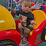 baby, child, toddler, kiddie_ride, toy_car, indoor, play_area, colorful, red, yellow, cartoon_mouse, seat_belt, footwear, shorts, tshirt, expression, steering_wheel, chairs, tables, carpet