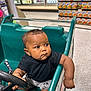 Zyon is registered to the contest to win money with this photo: baby, child, shopping_cart, supermarket, grocery_store, steering_wheel, curious, casual, black_shirt, shorts, floor, aisle, products, shelf, cart, infant, indoor, person, young_child, cute