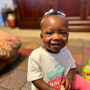 Amani is registered to the contest to win money with this photo: toddler, smiling, child, indoor, white_tshirt, pink_pants, beech_nut, smoothie_pouch, bow, hair_accessory, carpet, living_room, furniture, happy, cute, sitting, person, portrait, baby, playful
