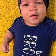 Kohlson is registered to the contest to win money with this photo: baby, infant, face, blue_eyes, hat, navy_blue_clothing, onesie, text_on_clothing, yellow_quilt, lying_down, child, cute, portrait, indoors, young_child, expression, small_hand, fabric_texture, closeup, person