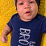baby, infant, face, blue_eyes, hat, navy_blue_clothing, onesie, text_on_clothing, yellow_quilt, lying_down, child, cute, portrait, indoors, young_child, expression, small_hand, fabric_texture, closeup, person