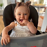 Nina a rejoint le concours — aidez-le/la à gagner de superbes lots ! baby, child, high_chair, headband, white_dress, smiling, happy, indoor, toy, furniture, person, cute, infant, face, hand, table, blurred_background, home, playful, portrait
