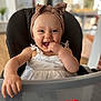 baby, child, high_chair, headband, white_dress, smiling, happy, indoor, toy, furniture, person, cute, infant, face, hand, table, blurred_background, home, playful, portrait