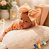 baby, blanket, bow, child, cozy, face, flowers, headband, indoor, infant, lying_down, nursery, peach_clothing, photography, pillow, portrait, smiling, soft_furnishings, vase, warm_lighting