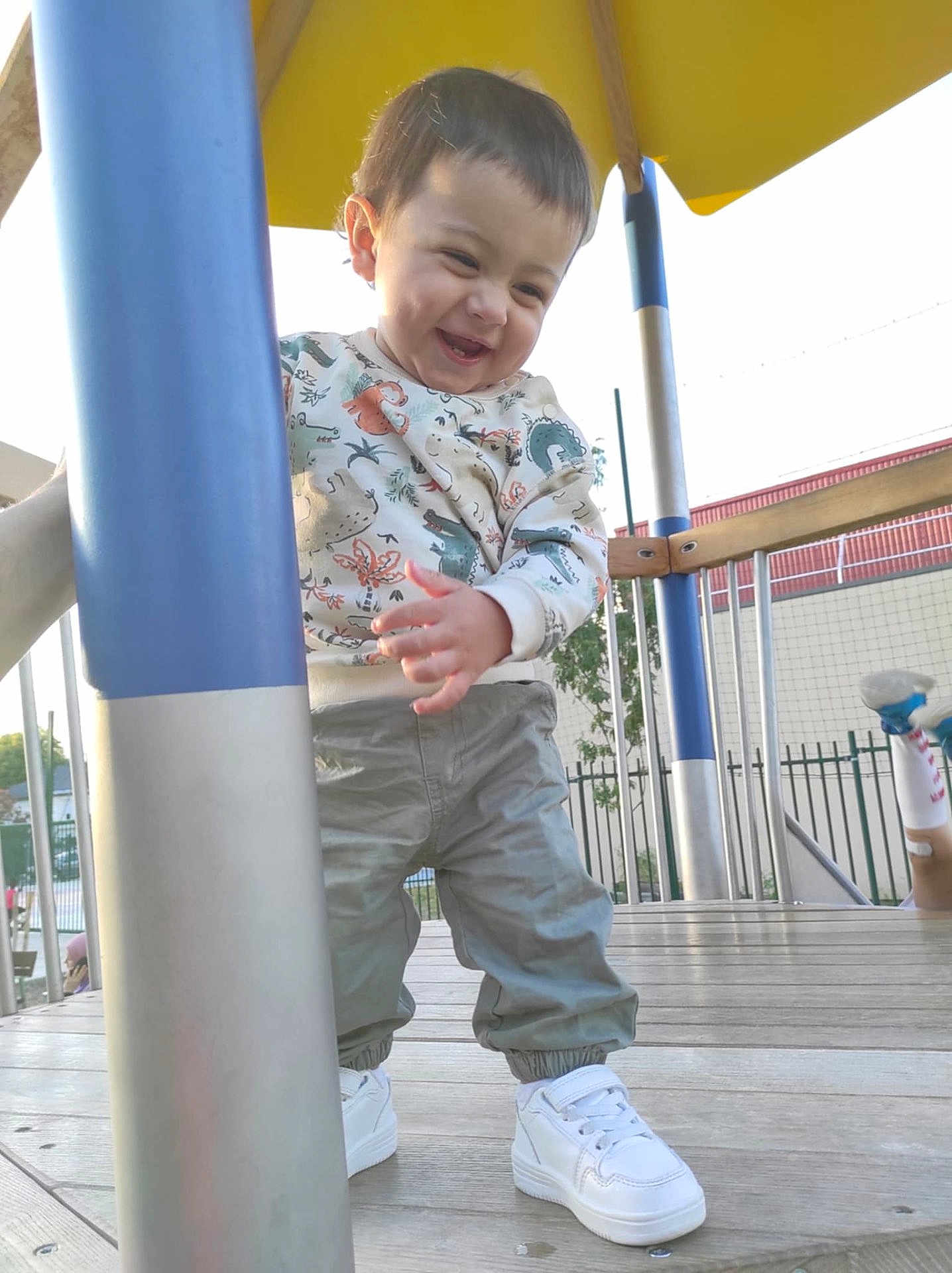 Ayden participe au concours pour gagner de l'argent avec cette photo : toddler, child, playground, outdoor, smiling, happy, sweater, pants, sneakers, pole, wooden_floor, yellow_canopy, metal_pole, fence, daylight, person, playing, casual_clothing, cute, fun