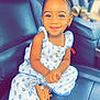 child, toddler, smiling, braided_hair, earrings, white_outfit, blue_flower_pattern, sitting, car_seat, leather_seat, footwear, sandals, portrait, indoor, happy, cute, young_child, person, fashion, casual