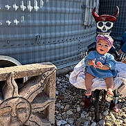 Amelia is registered to the contest to win money with this photo: baby, child, blue_dress, headband, skeleton_figure, red_horns, stone_sculpture, sun_face, rocks, outdoor, shadow_pattern, metal_wall, smiling, person, feet, clothing, playful, decor, daylight, happy
