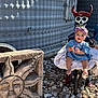 baby, child, blue_dress, headband, skeleton_figure, red_horns, stone_sculpture, sun_face, rocks, outdoor, shadow_pattern, metal_wall, smiling, person, feet, clothing, playful, decor, daylight, happy