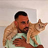 animal, beard, cat, cats, companionship, ears, fur, ginger_cat, green_shirt, holding, home, indoors, man, orange_tabby, person, pet, portrait, sitting, whiskers, white_wall