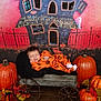 baby, sleeping, orange_clothing, pumpkin_pattern, wagon, metal_wagon, halloween_theme, pumpkins, autumn_leaves, wooden_floor, background_painting, haunted_house, smiling_baby, infant, holiday, fall_decor, festive, child, costume, cozy