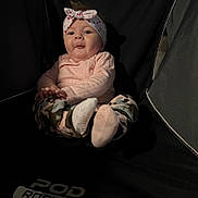 Willow is registered to the contest to win money with this photo: baby, headband, camo_pants, socks, pink_shirt, face, expression, indoor, chair, black_background, child, person, cute, sitting, clothing, footwear, infant, funny_face, headwear, podiatric