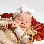 Romy a rejoint le concours — aidez-le/la à gagner de superbes lots ! baby, infant, newborn, sleeping, headband, bow, sparkly_dress, gold_dress, basket, wicker, blanket, plush, rust_blanket, studio_shot, white_background, portrait, hands, closeup, peaceful, cute
