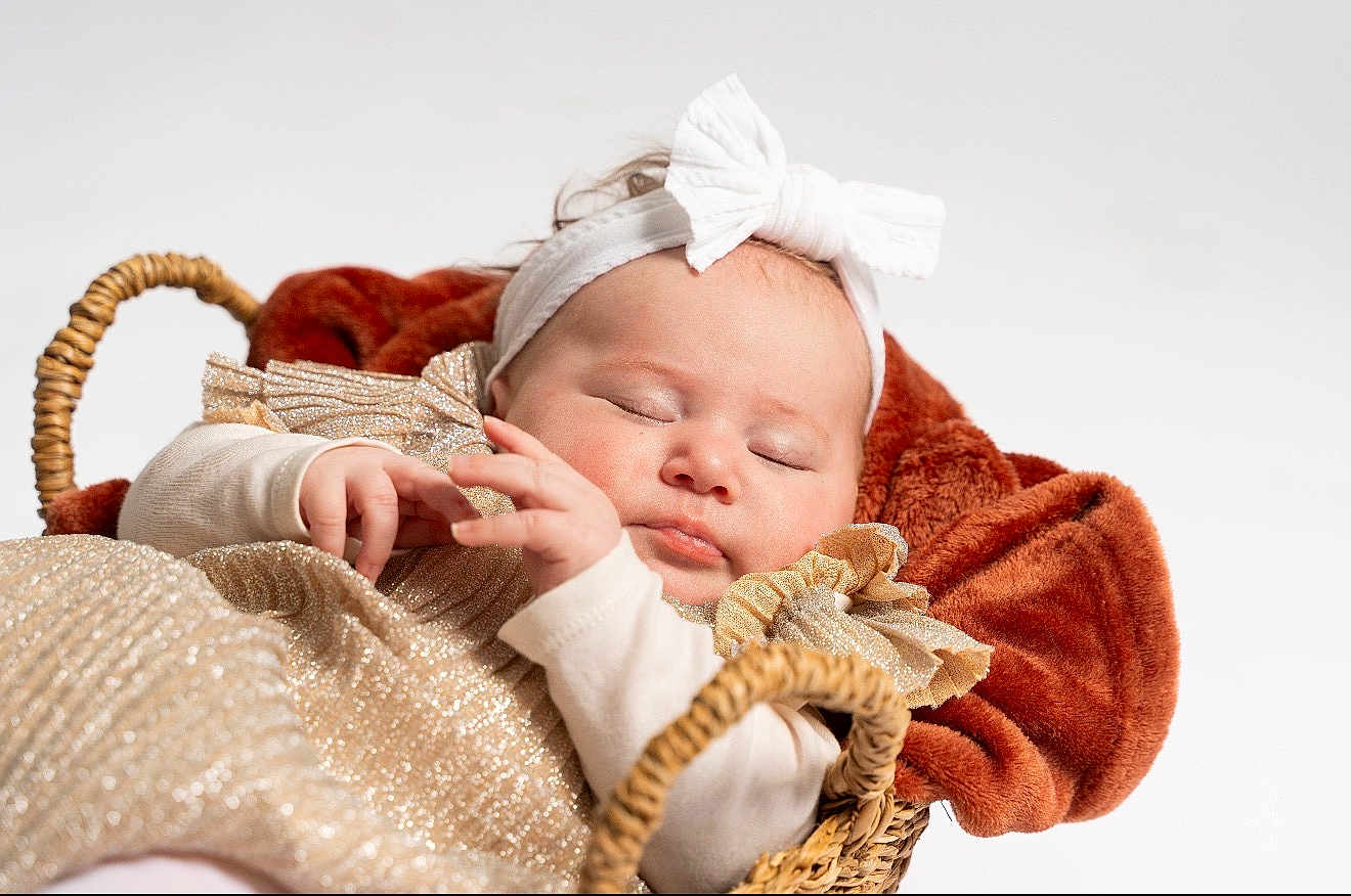 Romy a rejoint le concours — aidez-le/la à gagner de superbes lots ! baby, infant, newborn, sleeping, headband, bow, sparkly_dress, gold_dress, basket, wicker, blanket, plush, rust_blanket, studio_shot, white_background, portrait, hands, closeup, peaceful, cute