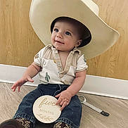 Henry is registered to the contest to win money with this photo: baby, child, cowboy_hat, boots, jeans, shirt, milestone_sign, floor, wooden_wall, smiling, sitting, indoor, person, cute, infant, portrait, hand, clothing, footwear, hat