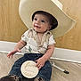 baby, child, cowboy_hat, boots, jeans, shirt, milestone_sign, floor, wooden_wall, smiling, sitting, indoor, person, cute, infant, portrait, hand, clothing, footwear, hat