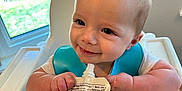 Henry is registered to the contest to win money with this photo: baby, child, high_chair, blue_bib, food_pouch, hand, window, greenery, indoor, feeding, smiling, happy, person, face, skin, table, plastic, nutrition, cute, young