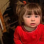 toddler, child, girl, red_clothing, indoors, portrait, face, brown_hair, pigtails, cute, young_child, seated, looking_at_camera, warm_lighting, casual, expression, innocent, person, close_up, soft_background