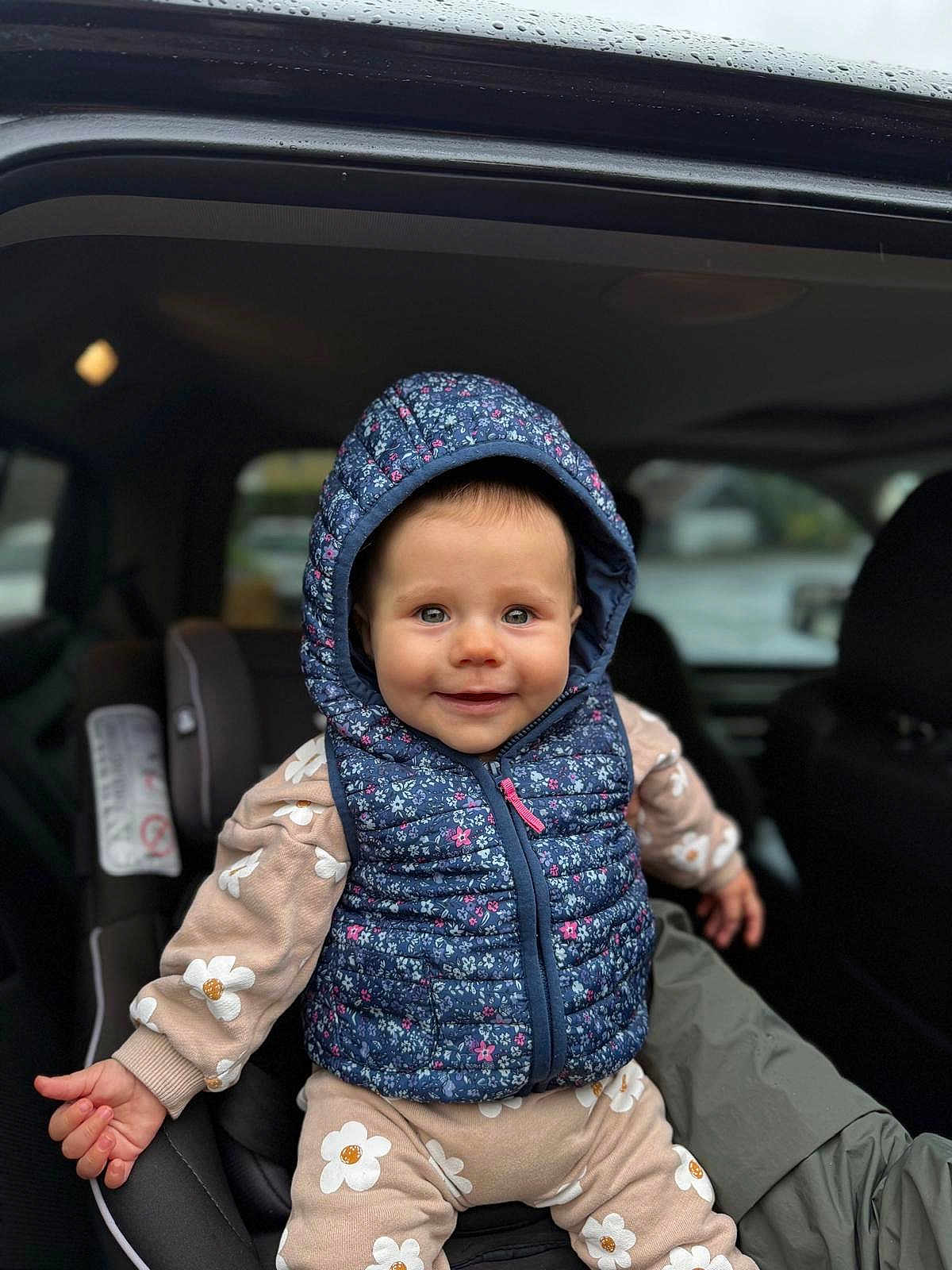 Railey participe au concours pour gagner de l'argent avec cette photo : baby, child, car_seat, hoodie, floral_pattern, smile, blue_hood, pink_zipper, seated, clothing, infant, cute, indoors, window, rain_drops, car_interior, person, young_child, happy, portrait