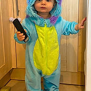 Hugo a rejoint le concours — aidez-le/la à gagner de superbes lots ! toddler, child, onesie, costume, monster_costume, blue, yellow, barefoot, remote_control, kitchen, tile_floor, wooden_cabinet, holding, standing, indoor, cute, baby, person, clothing, hood