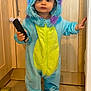toddler, child, onesie, costume, monster_costume, blue, yellow, barefoot, remote_control, kitchen, tile_floor, wooden_cabinet, holding, standing, indoor, cute, baby, person, clothing, hood