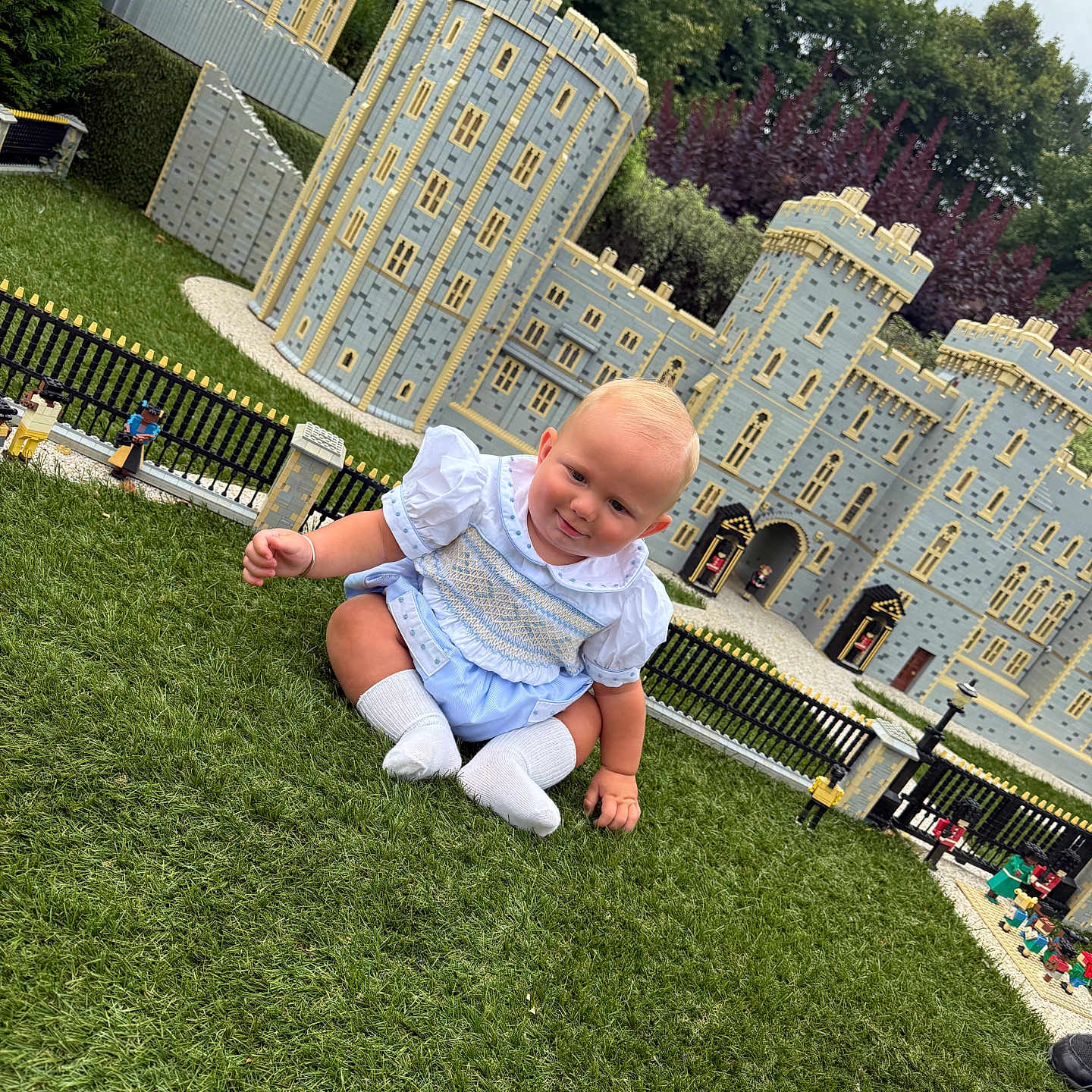 Tobias-John joined the competition — help win amazing prizes! baby, blue_outfit, castle, child, curious, cute, daylight, detailed, grass, greenery, lego, miniature_castle, nature, outdoor, park, playful, sitting, smiling, toy_figures, white_socks