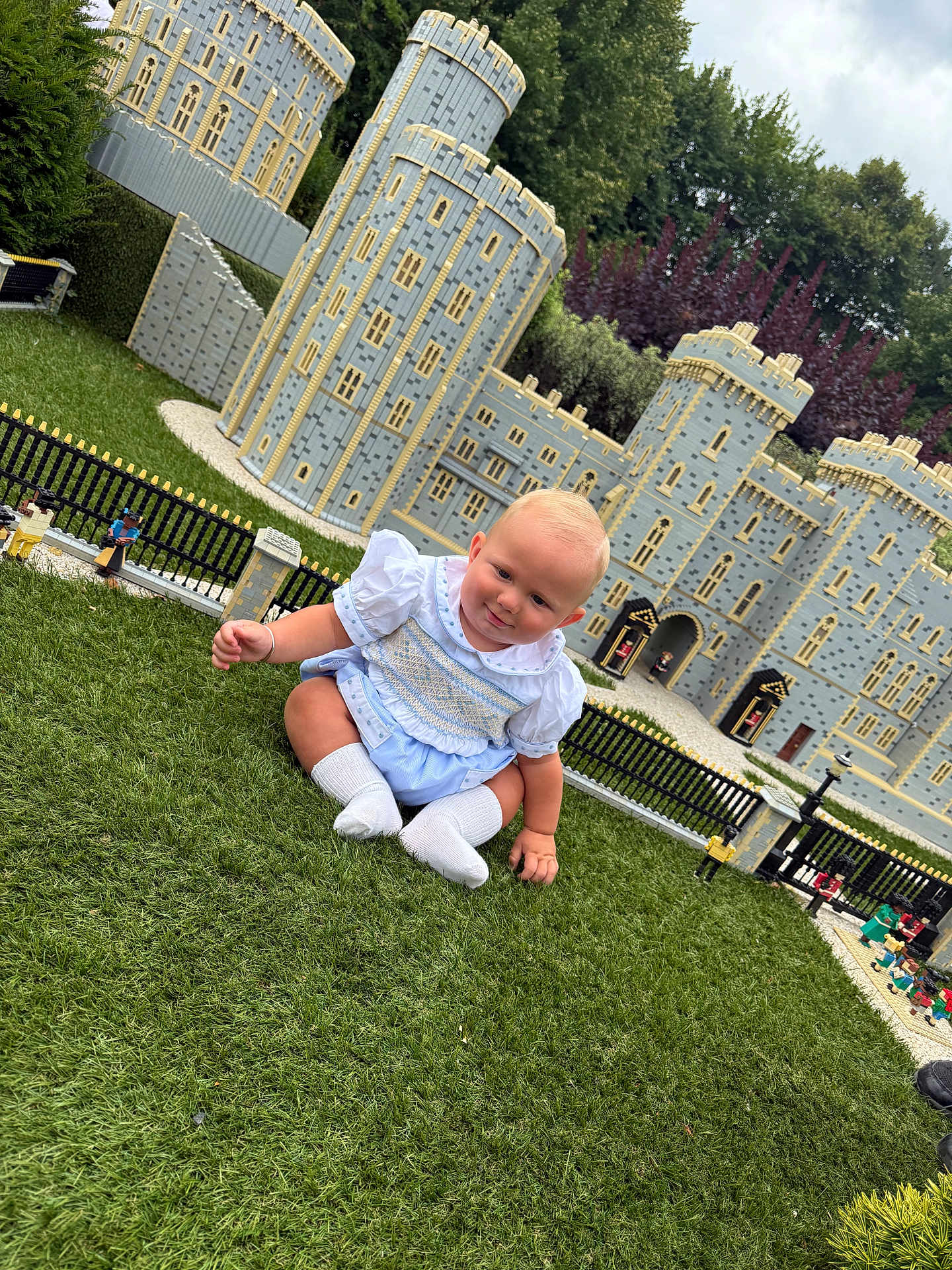 Tobias-John joined the competition — help win amazing prizes! baby, child, grass, miniature_castle, lego, outdoor, greenery, toy_figures, blue_outfit, white_socks, cute, smiling, playful, detailed, castle, park, daylight, nature, curious, sitting