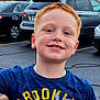 child, boy, red_hair, freckles, smile, blue_shirt, parking_lot, cars, outdoor, casual_clothing, happy, face, person, portrait, daylight, people, vehicle, asphalt, road, street