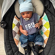 Karson is registered to the contest to win money with this photo: baby, infant, child, blue_hat, bib, pacifier, baby_rocker, cushion, colorful_pattern, jeans, black_sneakers, indoor, flooring, cute, seated, clothing, portrait, young_child, comfort, looking