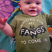 Sterling is registered to the contest to win money with this photo: baby, child, smiling, green_shirt, bat_graphic, text_on_clothing, play_mat, colorful_background, infant, cute, lying_down, happy, person, face, head, eyes, mouth, arm, hand, toddler