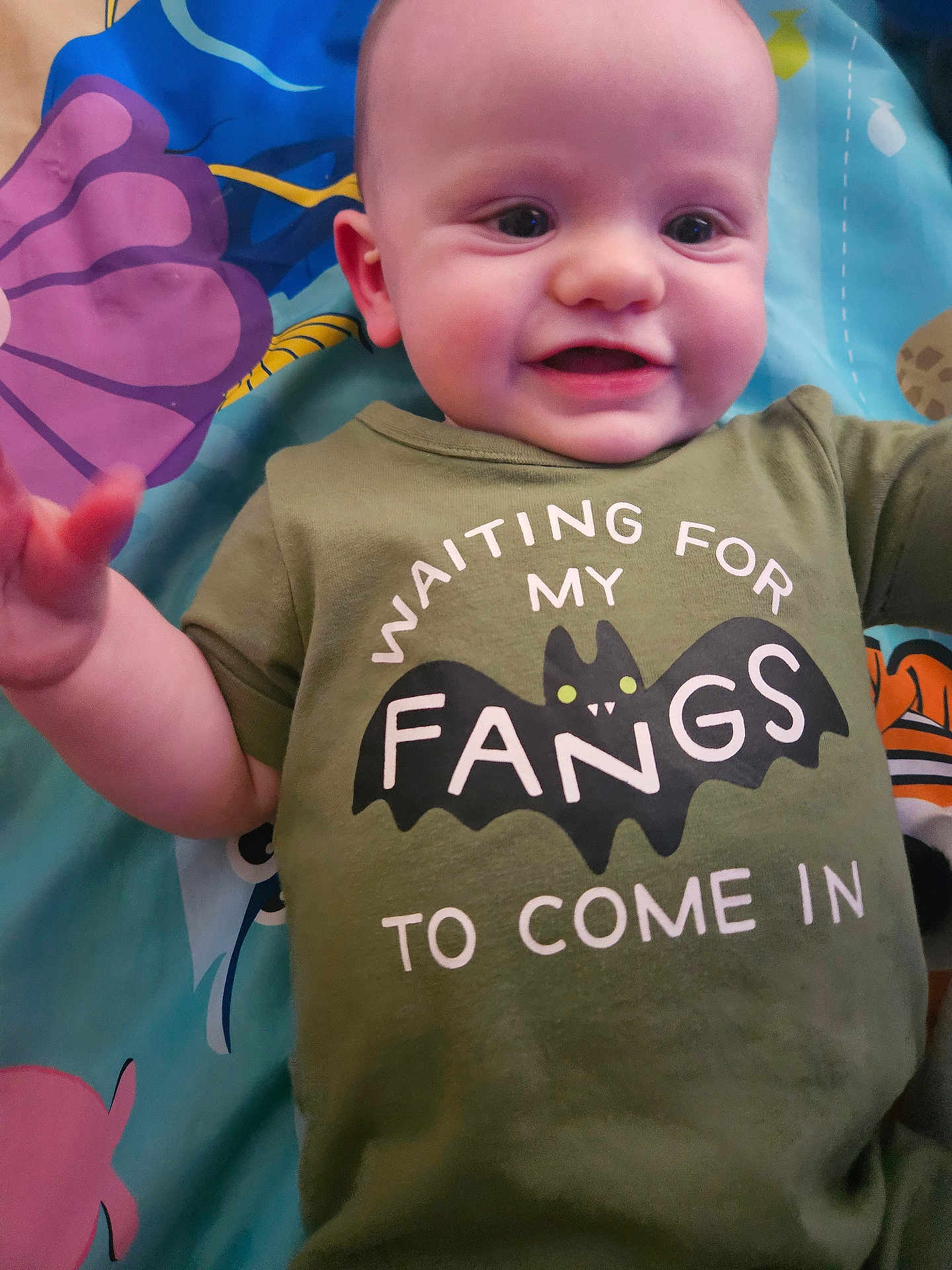 Sterling is registered to the contest to win money with this photo: baby, child, smiling, green_shirt, bat_graphic, text_on_clothing, play_mat, colorful_background, infant, cute, lying_down, happy, person, face, head, eyes, mouth, arm, hand, toddler