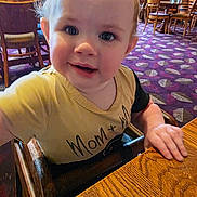 Lilly-Marie joined the competition — help win amazing prizes! toddler, child, high_chair, smiling, indoor, restaurant, wooden_chair, table, carpet, person, face, curly_hair, clothing, baby, hand, wood, background, eyes, expression, casual