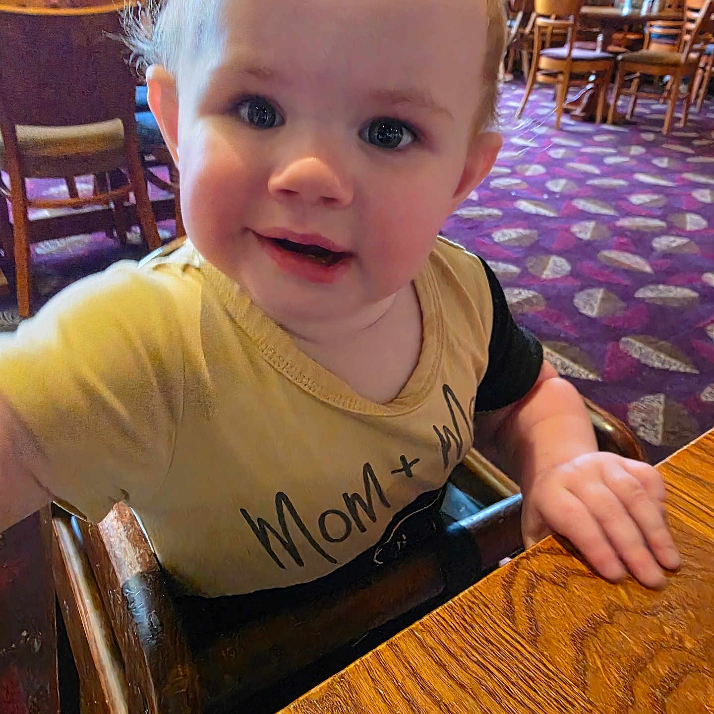 Lilly-Marie joined the competition — help win amazing prizes! baby, background, carpet, casual, child, clothing, curly_hair, expression, eyes, face, hand, high_chair, indoor, person, restaurant, smiling, table, toddler, wood, wooden_chair