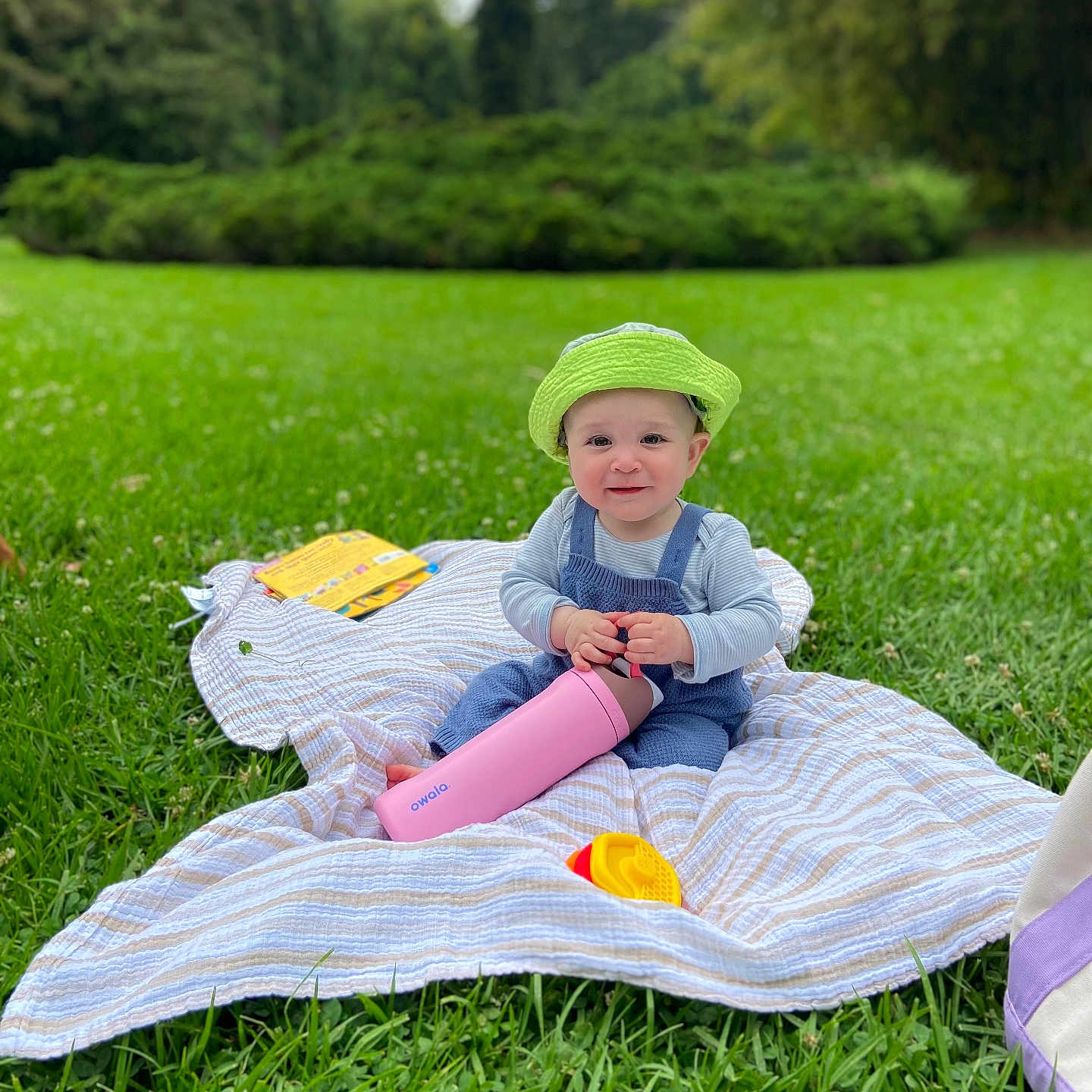 Isko joined the competition — help win amazing prizes! baby, child, green_hat, blanket, grass, outdoor, park, bottle, toy, smile, happy, nature, person, blue_overalls, striped_shirt, playtime, cute, daylight, sitting, relaxation