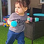 child, toddler, holding_ball, socks, ponytail, t_shirt, text_on_shirt, wicker_table, artificial_grass, colorful_balls, toy_blocks, patio, american_flag, window, glass_door, cushion, outdoor, play_area, portrait, standing