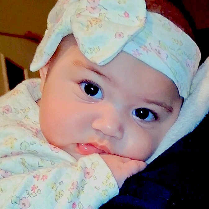 Ruth is registered to the contest to win money with this photo: baby, infant, face, headband, bow, clothing, floral_pattern, cute, closeup, resting, hand, soft_surface, portrait, child, young, indoor, person, skin, eyes, expression