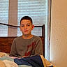 bed, blanket, boy, casual_clothing, child, curtain, face, home, indoor, natural_light, nike, person, portrait, quilt, relaxed, room, seated, sweatshirt, window, young