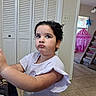 Monserrat is registered to the contest to win money with this photo: child, girl, curly_hair, indoor, pout, earrings, white_shirt, pink_tent, playroom, toy_slide, tile_floor, home, furniture, window, curtain, door, light_switch, person, portrait, expression