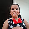 child, girl, polka_dot, red_bow, black_dress, earrings, curly_hair, smile, playful_expression, portrait, indoor, hand, bracelet, fashion, cute, young, person, face, happy, casual