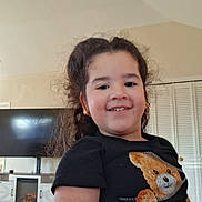 Monserrat is registered to the contest to win money with this photo: child, smile, curly_hair, black_shirt, teddy_bear, indoor, living_room, television, fireplace, earrings, cute, portrait, happy, home, wood_floor, furniture, casual_clothing, person, young_child, cozy