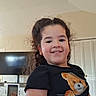 child, smile, curly_hair, black_shirt, teddy_bear, indoor, living_room, television, fireplace, earrings, cute, portrait, happy, home, wood_floor, furniture, casual_clothing, person, young_child, cozy