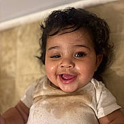 Leilani joined the competition — help win amazing prizes! baby, infant, child, smiling, curly_hair, drooling, stained_shirt, onesie, closeup, portrait, happy, chubby_cheeks, eyes, mouth_open, tongue, lying_down, floor, tiles, indoor, cute