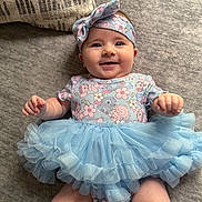 Aubree is registered to the contest to win money with this photo: baby, smiling, tutu, headband, floral, onesie, cute, infant, lying_down, soft_surface, pillow, happy, child, toddler, clothing, fabric, blue, pink, portrait, indoor