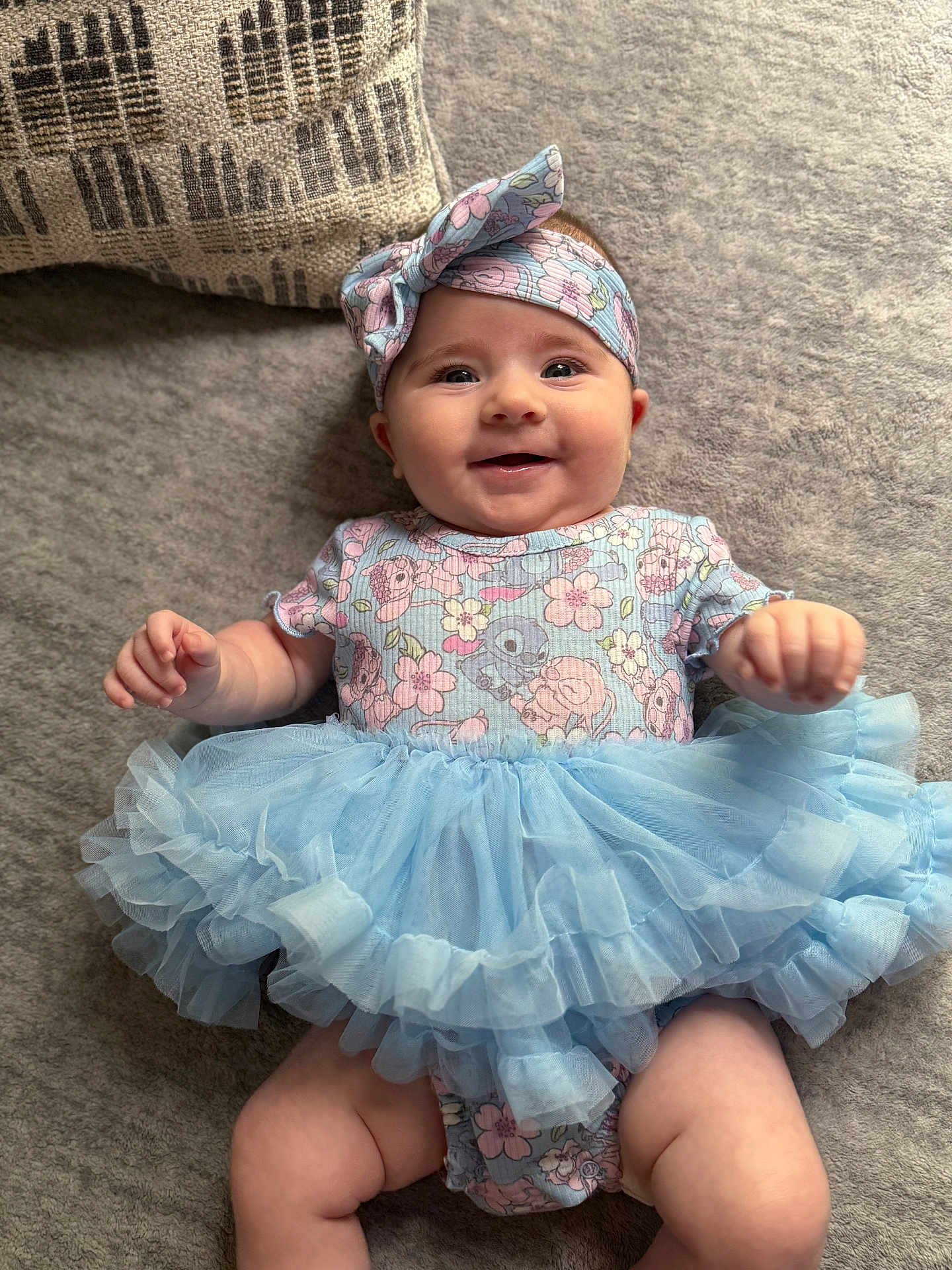 Aubree is registered to the contest to win money with this photo: baby, smiling, tutu, headband, floral, onesie, cute, infant, lying_down, soft_surface, pillow, happy, child, toddler, clothing, fabric, blue, pink, portrait, indoor