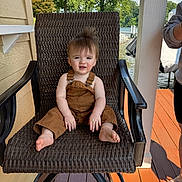 Kaizer is registered to the contest to win money with this photo: toddler, child, brown_overalls, chair, woven_chair, porch, outdoor, lake, trees, sunny, smile, barefoot, wooden_floor, person, hair, face, daylight, nature, relaxing, summer