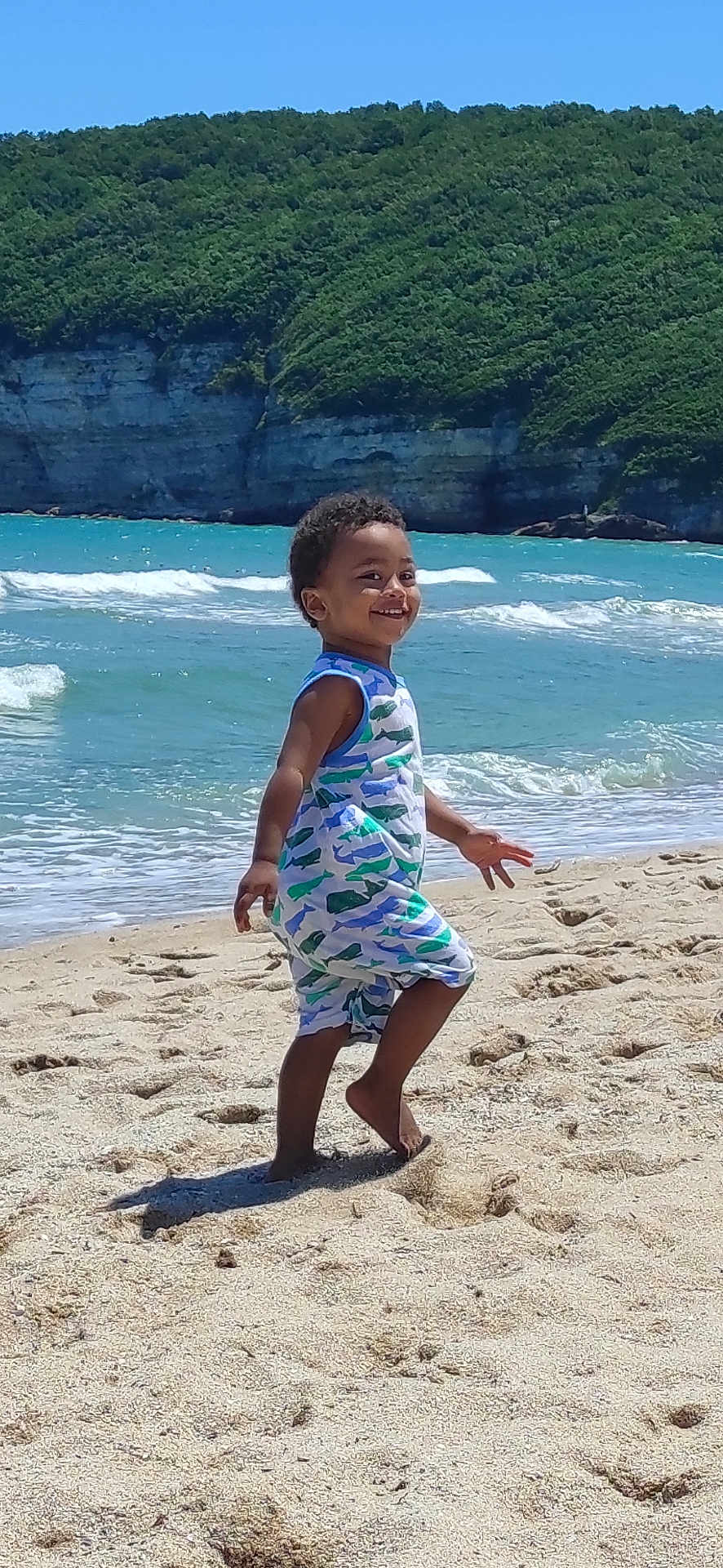 İbrahim joined the competition — help win amazing prizes! child, toddler, smiling, beach, sand, ocean, waves, cliff, greenery, blue_sky, sunlight, summer, vacation, barefoot, romper, portrait, playing, seashore, footprints, outdoor