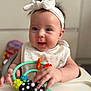 baby, headband, bow, high_chair, toy, hand, face, smile, infant, clothing, indoor, person, cute, child, play, colorful, expression, happy, closeup, young