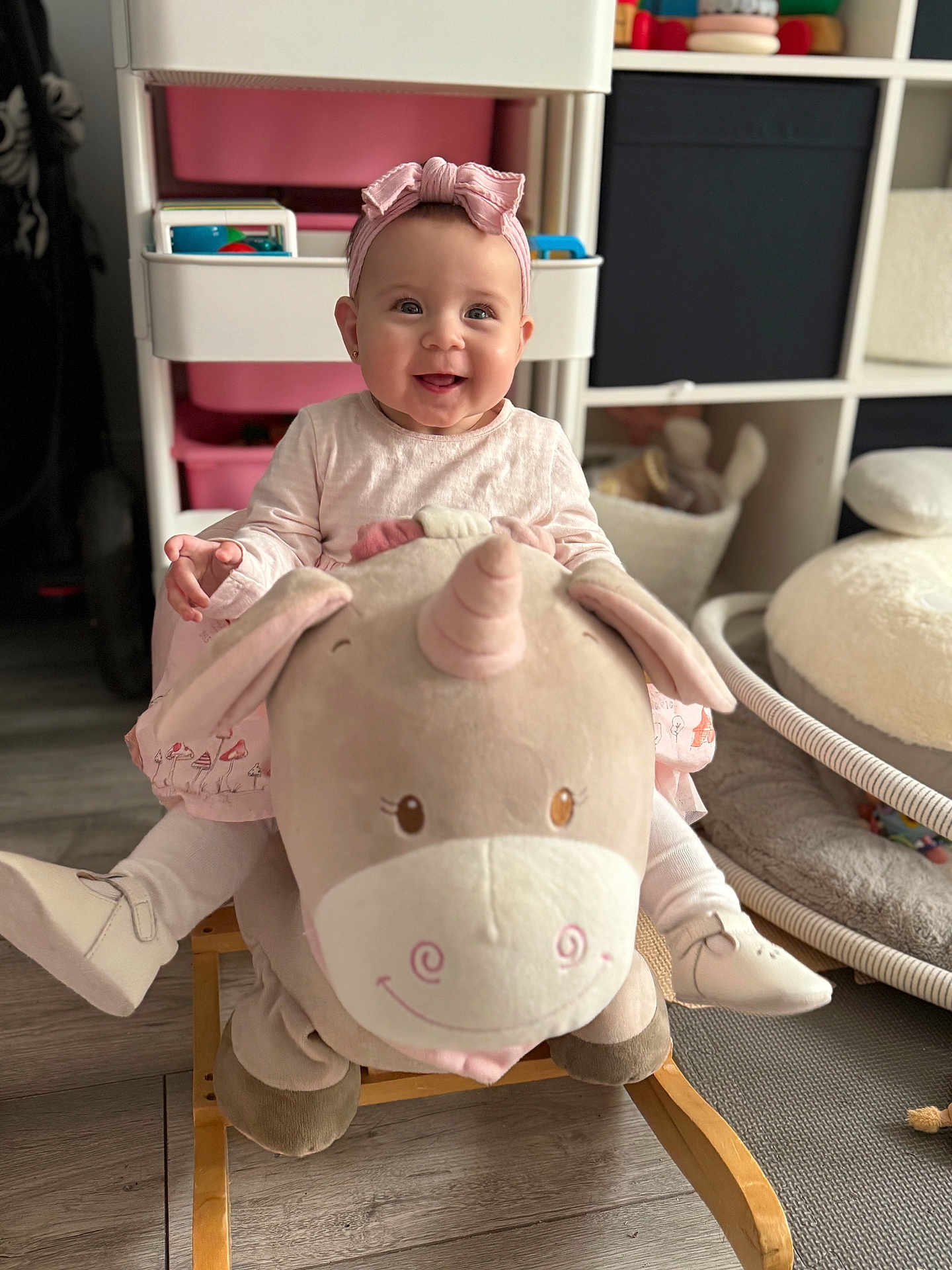 Emilia is registered to the contest to win money with this photo: baby, child, smiling, pink_headband, plush_toy, unicorn, rocker, indoor, playroom, toy_storage, wooden_floor, sitting, clothing, footwear, cute, happy, person, head, face, furniture