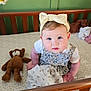 baby, infant, crib, headband, bow, blanket, teddy_bear, wooden_crib, blue_eyes, white_shirt, floral_dress, patterned_mattress, cute, child, indoors, soft_toy, baby_girl, sitting, looking_up, cozy