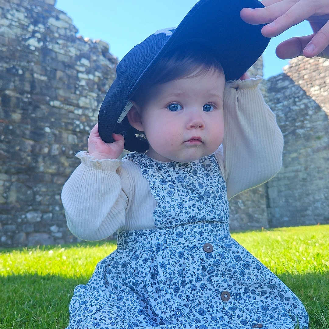 Lucia joined the competition — help win amazing prizes! baby, blue_sky, cap, child, curious_expression, daylight, dress, flower_pattern, foot, grass, greenery, hand, long_sleeve_shirt, nature, outdoor, person, playful, sitting, stone_wall, sunlight